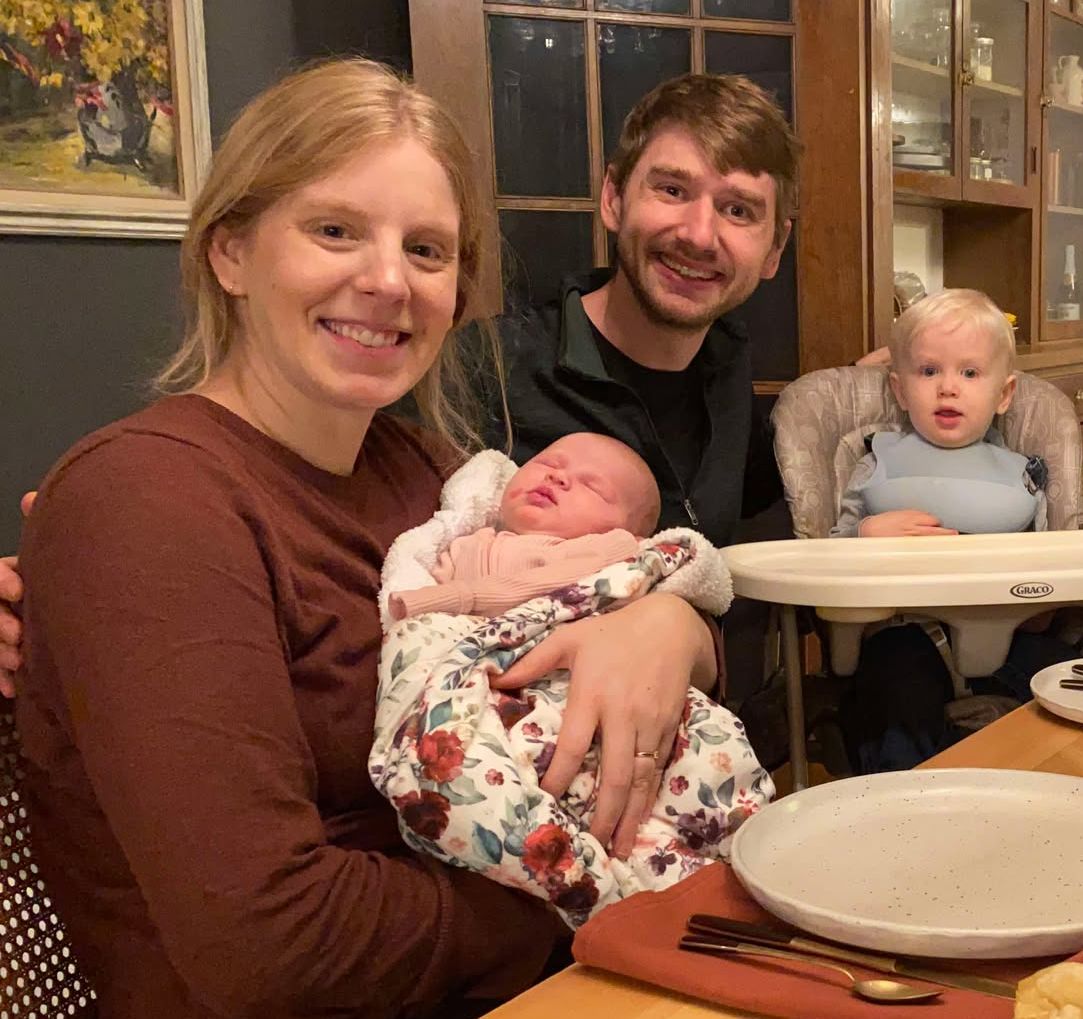 Photo from the 2024 Christmas letter: a woman holding a newborn baby at a dinner table, with a man and a toddler beside her